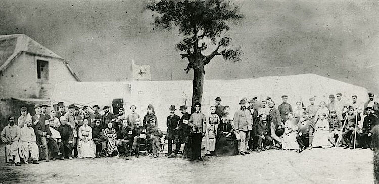 Patienten, Helferinnen und Helfer eines Kriegslazaretts, über dem die Fahne mit dem Roten Kreuz weht (DRK)