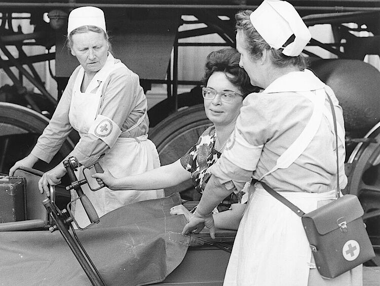 DRK der DDR: Bahnhofsdienst. Hilfe beim Umsteigen auf dem Bahnsteig für eine Frau im Rollstuhl, vermutlich in Dresden